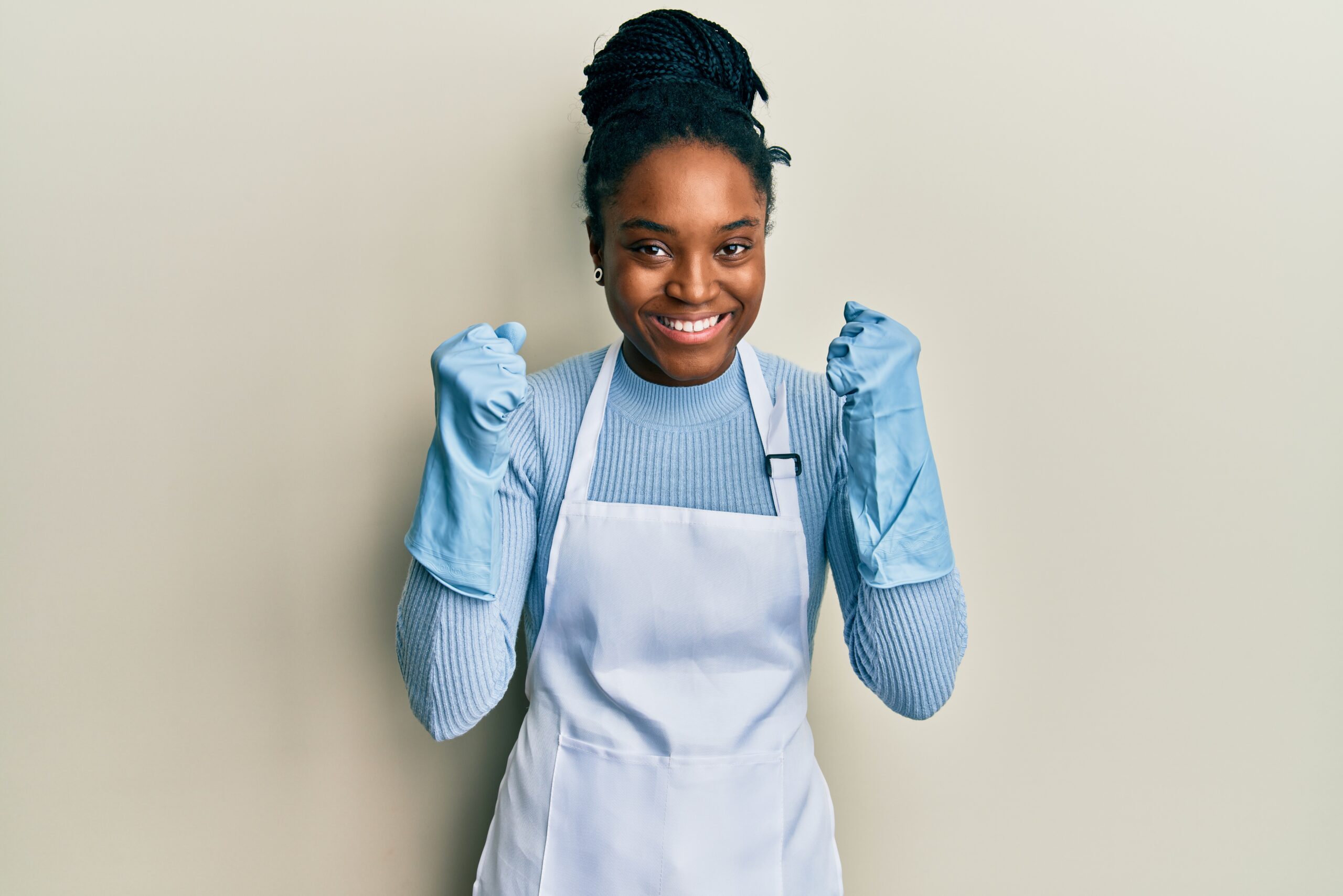 African american woman with braided hair wearing cleaner apron and gloves celebrating surprised and amazed for success with arms raised and open eyes. winner concept. African american woman with braided hair wearing cleaner apron and gloves celebrating surprised and amazed for success with arms raised and open eyes. winner concept.